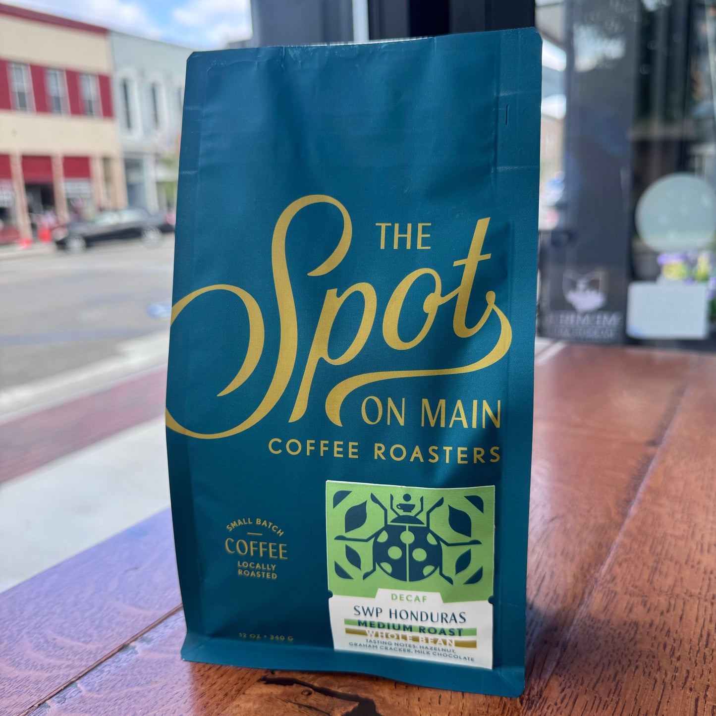 A bag of 'The Spot On Main' coffee roasters, specifically SWP Honduras Decaf, standing on a wooden table in front of a window.